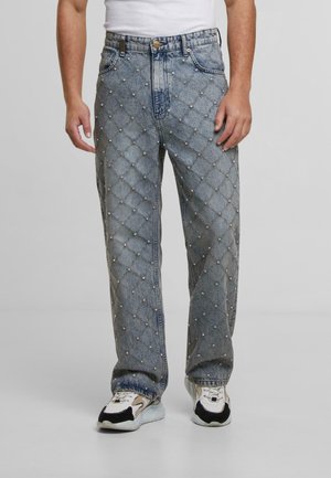 Man in lichtblauwe spijkerbroek met een gequilt patroon en kleine studs, gecombineerd met witte sneakers en een wit shirt.