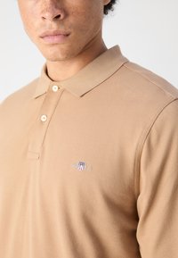 Camisa polo beige hecha de tela texturizada, con una tapeta de tres botones y un logo bordado en el lado izquierdo del pecho.