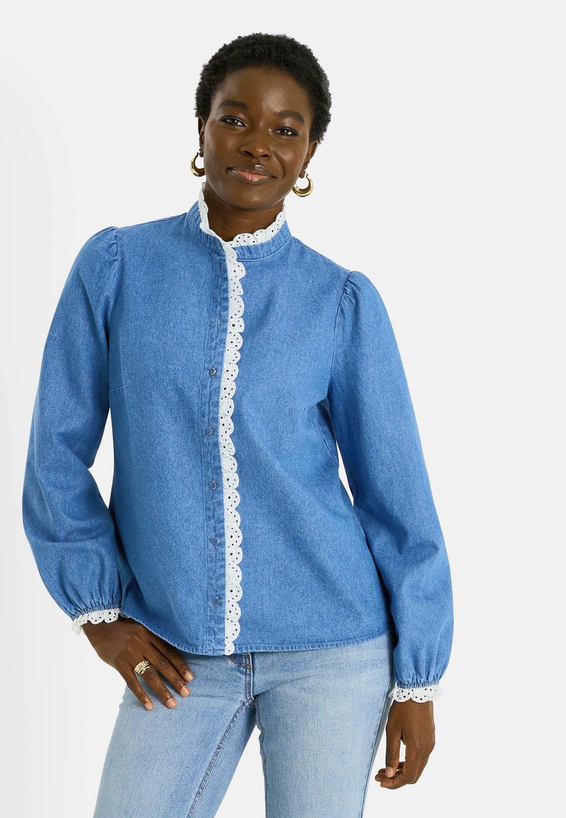 Camisa de mezclilla azul con mangas abullonadas y borde de encaje blanco a lo largo de la tapeta frontal y los puños. Los botones son visibles en el centro.