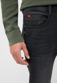 Męska dłoń spoczywająca blisko kieszeni czarnych dżinsów z małą czerwoną metką, w zielonej koszuli z długim rękawem o wzorze wafelowym.