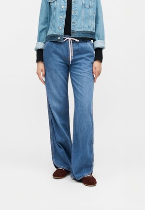 Person iført en lyseblå denimjakke med opkrogede ærmer, sort top, blå vidde jeans med snøre i taljen og brune sko.