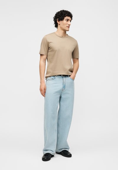 Νέος άντρας στέκεται με το χέρι στην τσέπη, φοράει μπεζ κοντομάνικο μπλουζάκι, ανοιχτό μπλε φαρδιά τζιν και μαύρα παπούτσια σε λευκό φόντο.