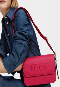 Bolso de hombro de cuero texturizado rosa con el logo "TOUS" en relieve, herrajes dorados y una correa roja contrastante. Cierre de solapa suave.