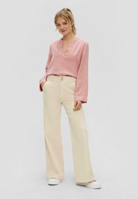 Lichtroze blouse met lange mouwen en een V-hals, met wijde mouwen, gecombineerd met crème kleurige, hoge taille, wijde broek en witte sneakers.