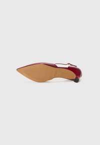 Escarpin en cuir verni rouge avec un bout pointu, une bride à la cheville réglable et une semelle en caoutchouc beige avec des lignes de grip texturées.