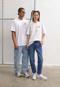 Two individuals stand side by side, each wearing white cotton t-shirts with a small multicolored graphic. The man has light blue jeans and sneakers, while the woman wears dark blue jeans and white sneakers.
