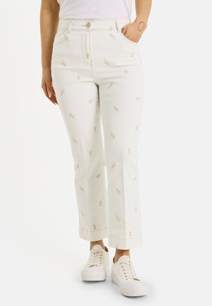 Pantalones blancos de corte recto con pequeños estampados florales beige, combinados con zapatillas blancas, llevados por una persona con una mano en el bolsillo.