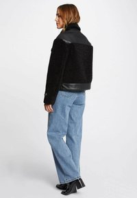 Giacca nera in pile e pelle con colletto alto, design cropped e maniche lunghe. Indossata con jeans azzurri chiari e stivaletti neri con tacco.