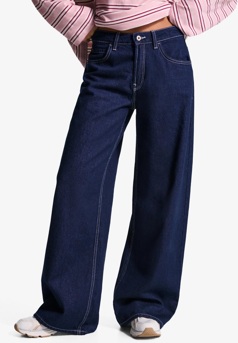 Široke temno jeans hlače z belimi šivi. Imajo klasičen pet-pocket dizajn in zaklep s trdnim pasom.