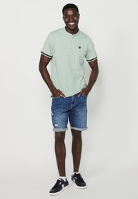 Polo verde chiaro a maniche corte con motivo sottile, rifiniture nere, abbinato a pantaloni shorts di jeans arrotolati e sneakers blu navy. Tessuto dalla consistenza liscia.