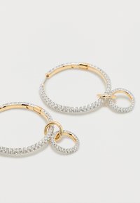 Σκουλαρίκια κρίκοι χρυσού με διαφανείς στρας, καθένα με δύο μικρότερους συνδεδεμένους κρίκους στρας που κρέμονται ως γοητεία, σε λευκό φόντο.