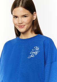 Niebieska bluza z miękkiego materiału, z okrągłym dekoltem i luźnym krojem. Z przodu znajduje się biała wyszywana fraza "Stay wonderful."