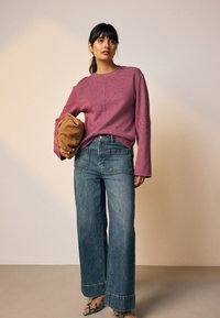 Pull rose en maille à manches longues ; jean denim bleu à jambes larges ; tenant un sac marron ; portant des chaussures à motifs ; fond simple.