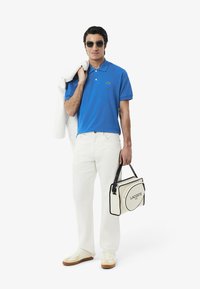 Blå polo med grön logotyp, vita jeans och vita sneakers med bruna sulor. Bär en krämfärgad väska med svarta detaljer och text.