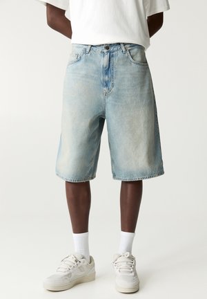 Homme portant un short en jean bleu clair arrivant aux genoux, des chaussettes blanches, des baskets blanches et un t-shirt blanc, sur un fond uni.