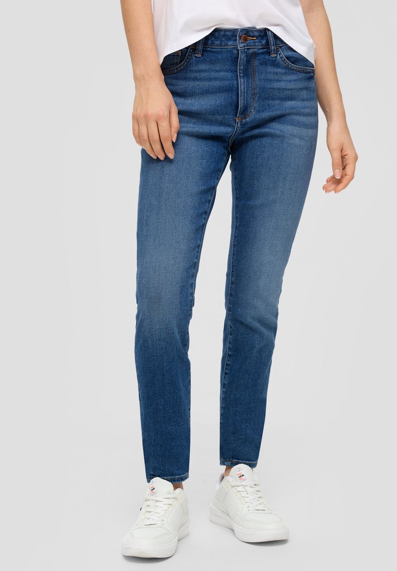 Jeans azul oscuro con una silueta ajustada, que presentan un diseño clásico de cinco bolsillos y un ligero desgaste. Combinados con zapatillas blancas.