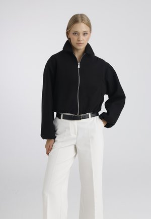 Femme blonde portant une veste noire zippée à col montant, un pantalon blanc taille haute et une ceinture noire, posant avec une main dans la poche.