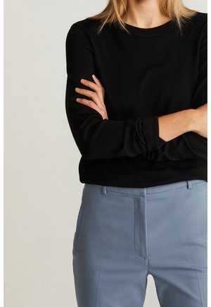 Personne portant un haut noir à manches longues et un pantalon bleu clair, debout les bras croisés devant un fond uni.