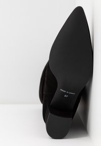 Czarny zamszowy but na wysokim obcasie z wydłużonym noskiem, charakteryzujący się gładką, teksturalną powierzchnią. Podeszwa oznaczona "Wyprodukowano w Hiszpanii" oraz rozmiar "37."