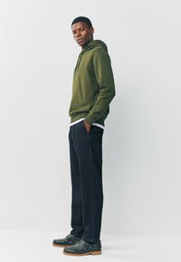 Olive green hoodie with a drawstring, worn over a white shirt; paired with dark trousers and black shoes with brown soles.