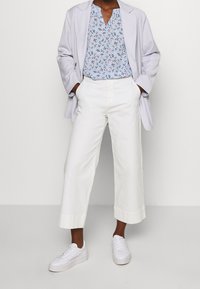 Hellblauer Blazer über einer blumigen blauen Bluse, kombiniert mit weißen, kürzeren Hosen und weißen Sneakers. Glatter Stoff, entspannte Passform.