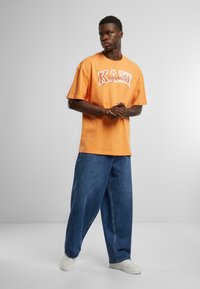 Orange bomuld t-shirt med "KANI Originator" grafik, parret med løse blå denim jeans og hvide sneakers, stående mod en neutral baggrund.