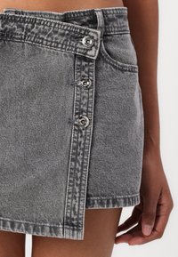Jupe en denim gris avec un ourlet effiloché, dotée de trois boutons métalliques sur le côté et d'une ceinture à motif distinct.
