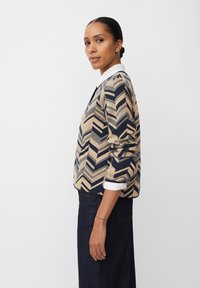 Giacca trapuntata con un motivo a zigzag nei toni navy e beige. Tessuto texture, scollo rotondo e maniche arrotolate con una camicia bianca sotto.