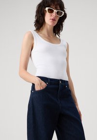 Weißes, geripptes Tanktop mit Rundhalsausschnitt, kombiniert mit dunkelblauen Jeans aus Denim mit Vorderentaschen und goldfarbener Hardware.