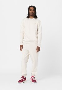 Crème-kleurige sweatshirt en bijpassende joggers, gemaakt van zacht materiaal. Heeft elastische manchetten en een klein logo op de borst. Draagt met bordeauxrode sneakers.