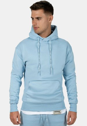 Jeune homme regardant à gauche portant un sweat à capuche bleu clair avec des cordons, une poche kangourou et un pantalon de survêtement assorti avec un maillot de corps blanc.