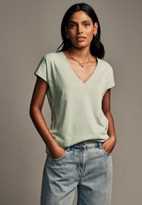 Minzgrünes V-Ausschnitt-T-Shirt aus weichem Stoff, lockere Passform, kurze Ärmel. Kombiniert mit hellblauen Jeans aus Denim mit vorderen Taschen.