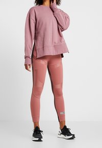 Žena oblečená do fialového oversized svetru a ladících práškově růžových sportovních legín s černobílými běžeckými botami, stojící na bílém pozadí.