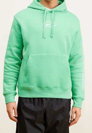 Homme portant un sweat à capuche Nike vert menthe avec poche avant, cordons et logo blanc, associé à un pantalon noir, sur un fond uni.