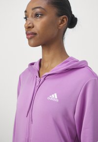 Fialová bunda na zips vyrobená z mäkkého materiálu, s bielym logom Adidas na ľavej hrudi a kapucňou so šnúrkami.