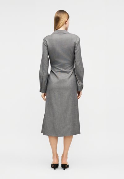 Γυναίκα στέκεται πλάτη, φορόντας γκρι midi φόρεμα με μακριά μανίκια και γιακά, και μαύρα παπούτσια με χαμηλό τακούνι, σε λευκό φόντο.