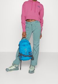 Felpa rosa tagliata, pantaloni color teal e uno zaino blu con tasche in rete. Le calzature presentano colori grigi e verdi con lacci.