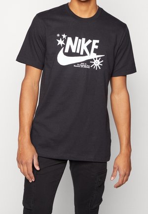 Osoba nosząca czarną koszulkę Nike z białym logo, gwiazdkami i grafiką słońca, w połączeniu z czarnymi spodniami typu cargo.