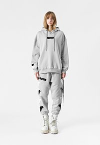Sudadera holgada de color gris claro con bolsillo frontal y cordones, combinada con pantalones jogger a juego que presentan detalles y parches en negro y blanco.