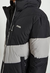 Giubbotto imbottito nero e grigio con chiusura a zip, cappuccio regolabile e tasche laterali. Il materiale è trapuntato e presenta un logo sul petto.
