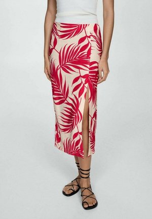 Mujer con una falda midi beige con estampado de hojas tropicales rojas y una abertura frontal, combinada con sandalias planas negras con tiras y una blusa blanca sin mangas.