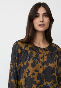 Blusa de manga larga en un patrón floral negro y mostaza, con un cuello redondo y detalles de botones en la parte frontal. Tela suave y texturizada.