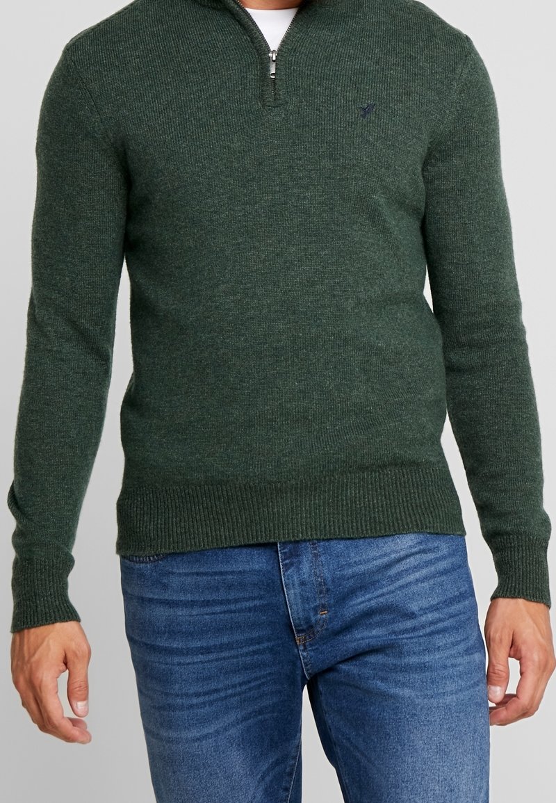 Uomo che indossa un maglione lavorato a maglia verde scuro con zip a quarti sopra una camicia bianca, abbinato a jeans blu, su uno sfondo neutro.