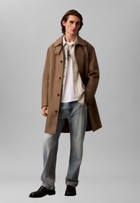 Brun trenchcoat med knappelukking foran, lagd over en hvit skjorte og en lys grå stripete skjorte, kombinert med lyseblå jeans og svarte sko.