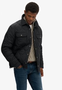 Gesteppte schwarze Jacke mit zwei Brusttaschen, silbernen Druckknöpfen und einem Kragen, darüber getragen ein beiger Strickpullover mit Zopfmuster.