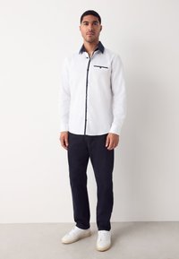 Camicia bianca a maniche lunghe con colletto e dettaglio tasca blu navy, abbinata a pantaloni blu navy e sneakers bianche, mostrata su uno sfondo neutro.