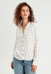 Chemise blanche à boutonnière avec un petit imprimé de coeurs noirs, à manches longues et col. Associée à un jean en denim bleu clair.