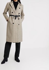 Double-breasted beige plaid trench coat with a black belt, featuring large buttons, a notched collar, and side pockets, paired with black pants.