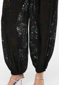 Czarne spodnie z koronki z kwiatowymi wzorami, o fakturowanym wykończeniu i marszczonymi nogawkami u dołu, zestawione z srebrnymi sandałami na wysokim obcasie.