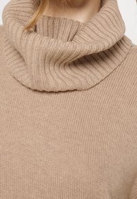 Beige gebreide sweater met een brede, geribbelde kraag. De textuur is zacht en glad, met een gelijkmatig patroon door de stof.